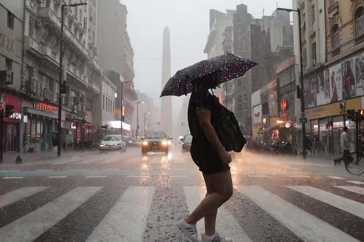 Una mujer caminando bajo la lluvia con el Obelisco en CABA de fondo.